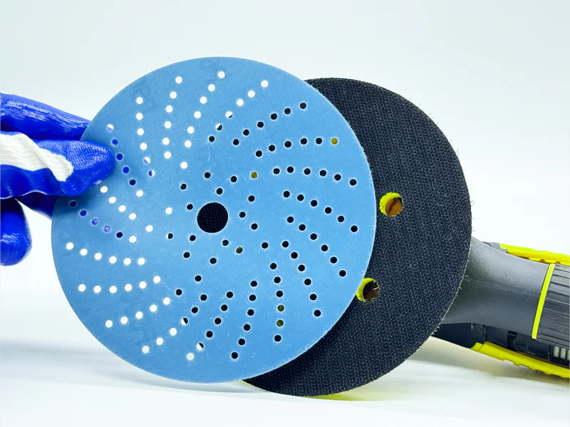 Blue zirconia alumina sanding disc with multiple holes, held by gloved hand in workshop setting
