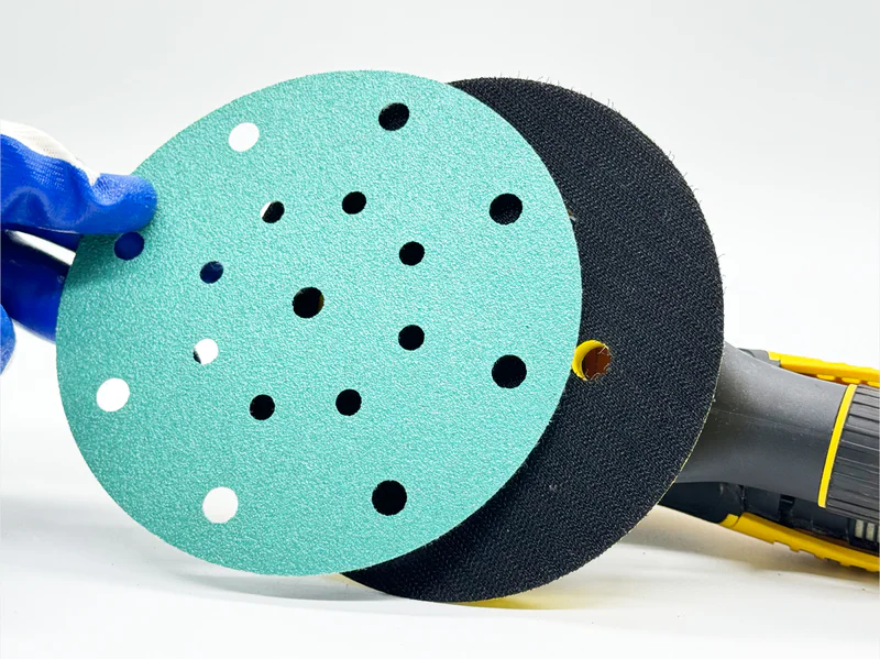Green zirconia sanding disc with multiple holes, held by gloved hand in workshop setting