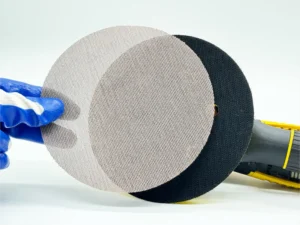 Gray mesh net sanding disc, aluminum oxide abrasive, held by gloved hand with sander in workshop setting