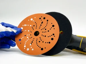 Orange ceramic sanding disc with multiple hole patterns held by gloved hand in workshop setting