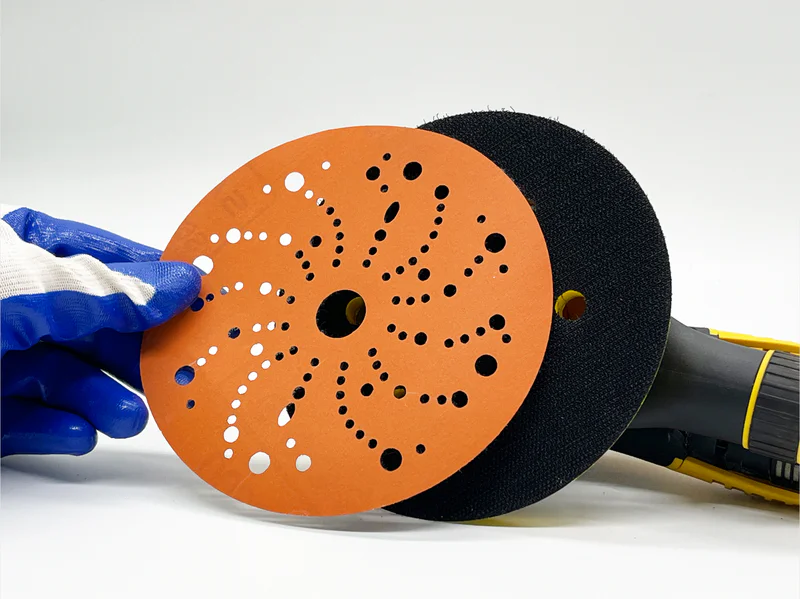 Orange ceramic sanding disc with multiple hole patterns held by gloved hand in workshop setting